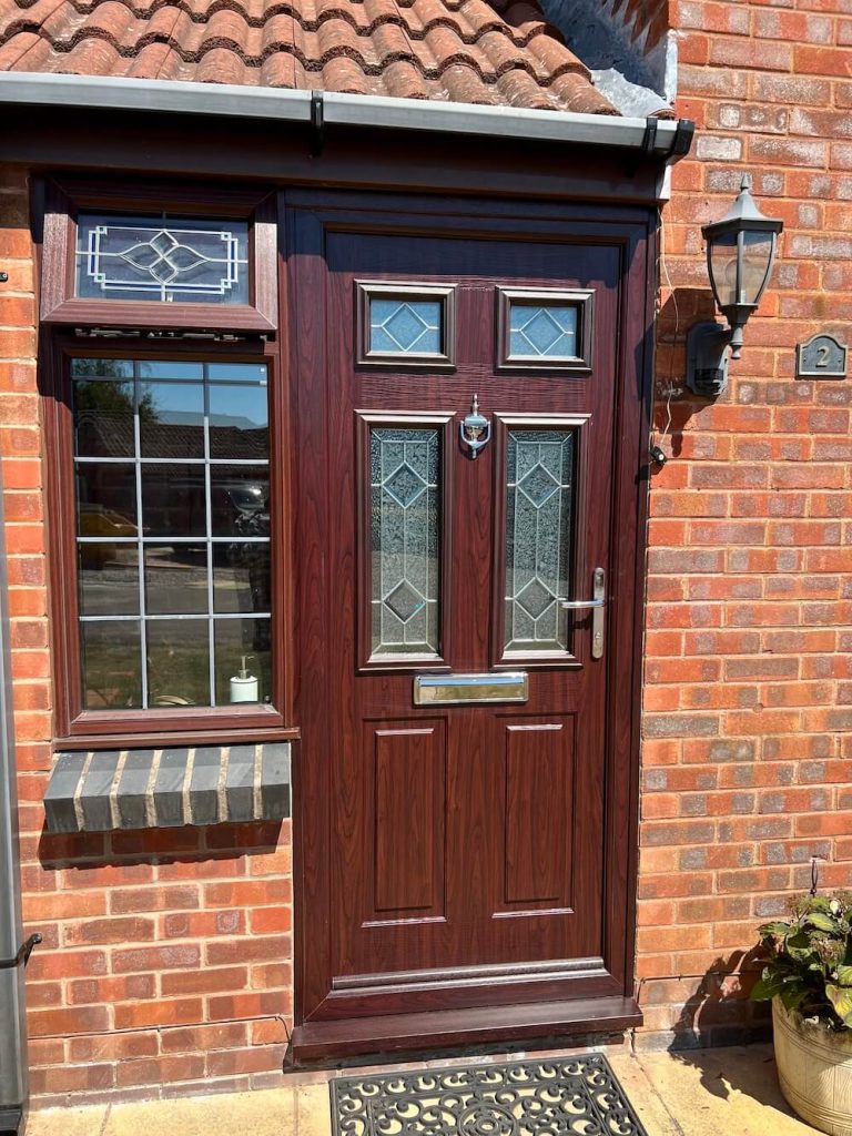 Dark wood-effect composite front door with stained glass panels, set in a red-brick house with a side window and lantern light.