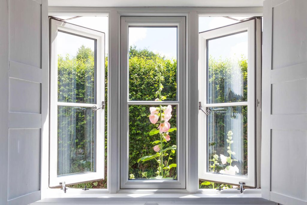Open double glazed windows with white frames, providing a clear view into a sunny garden with lush greenery and blooming pink flowers, in a room with white walls and a visible silver handle and latch on the window.