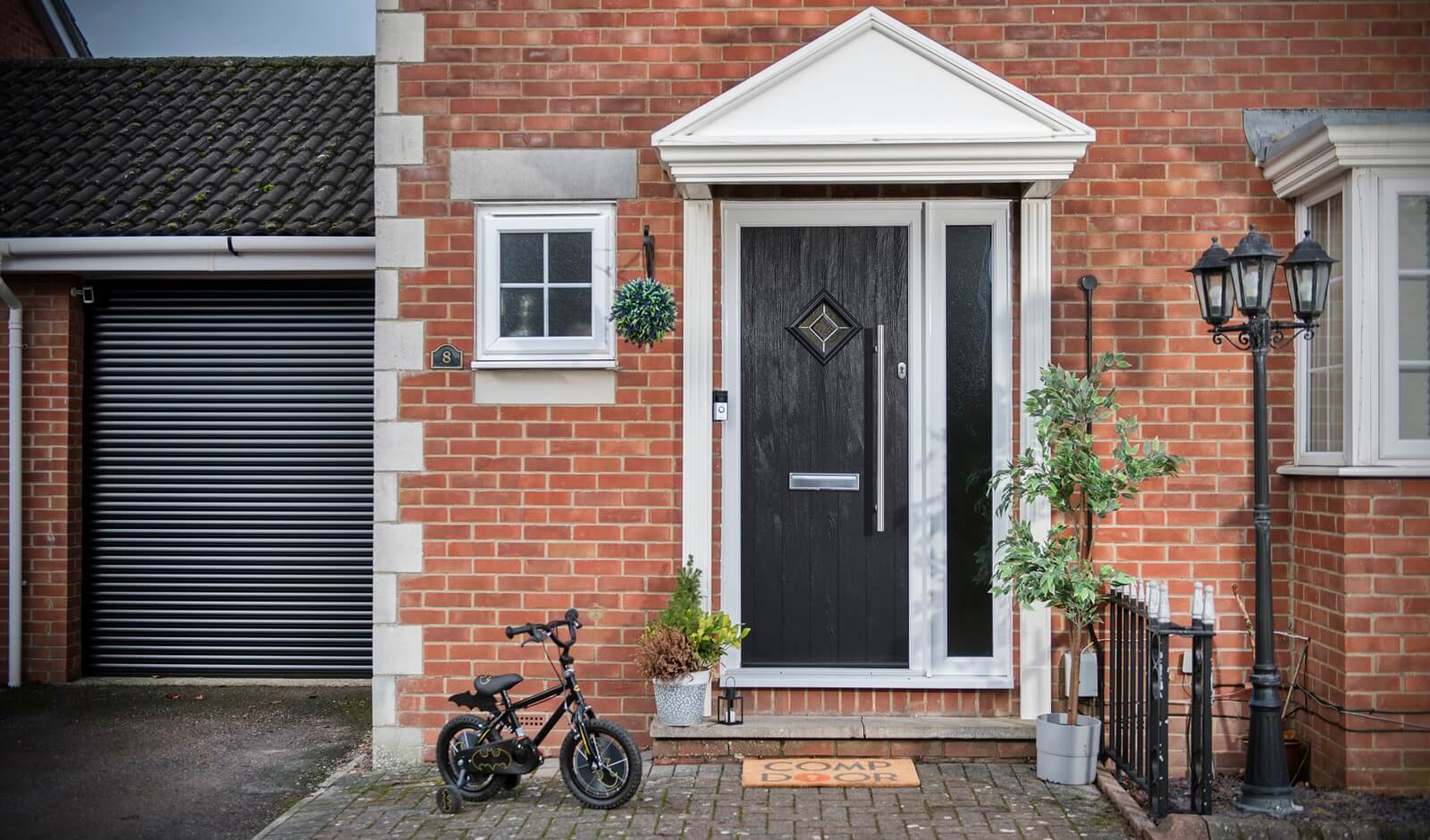 This image presents a sophisticated black composite door, beautifully offset by elegant silver hardware. Notably, the sleek letterbox is thoughtfully situated halfway up the door, while a secure lock with a matching pole handle, extending half the door's length, is centrally placed on the right side. Enhancing the door's appeal, there is a striking full-length glass panel, taking up a fourth of the door's width, alongside a patterned glass panel positioned towards the top at head height, both featuring an eye-catching diamond design. The combination of these modern design elements and practical features creates a door that is both inviting and secure. Complementing the entrance is a 'Comp Door' doormat, neatly placed outside, adding a welcoming touch to this elegant entryway.