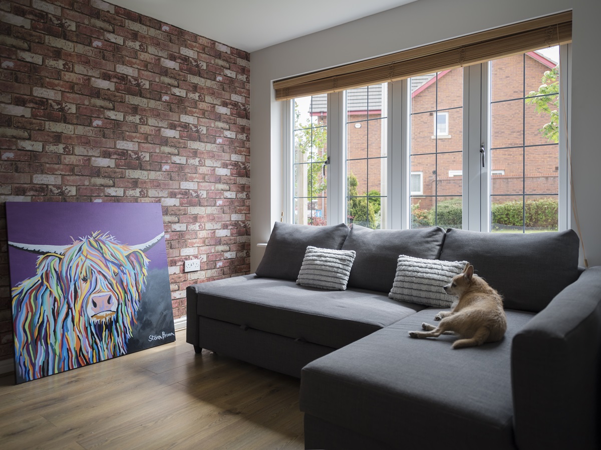 A stylish urban living room with a large Residence 7 timber effect window, dark grey sofa, brick accent wall, and a colorful contemporary painting, with a dog resting peacefully.