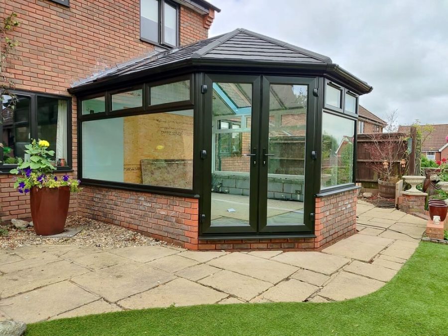 A modern conservatory with a black tiled roof, large glass windows, and a brick base, attached to a traditional home. The design highlights energy efficiency and contemporary style.