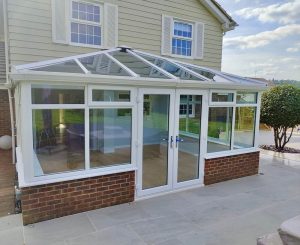 A modern conservatory with a pitched glass roof, white uPVC frames, and a brick base, designed for natural light and seamless views of a landscaped garden. The image highlights a versatile and stylish extension for any home.
