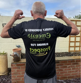 iew from behind of a runner, Nigel, flexing his arms while wearing a black running shirt sponsored by Emerald and running for Prospect Hospice