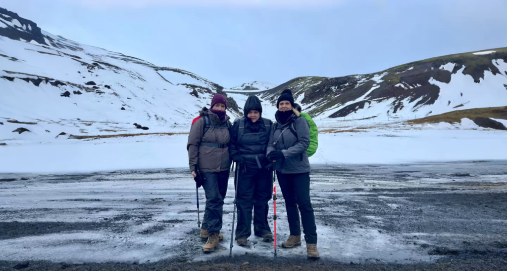 Three people dressed in heavy winter hiking gear, including hats, jackets, and backpacks, are standing on an icy path with snow-covered mountains and a cloudy sky in the background. They are all holding hiking poles.