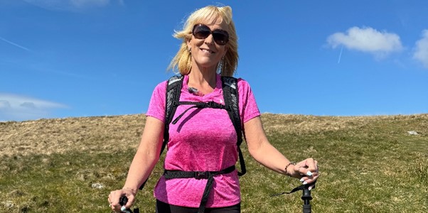 A smiling woman with blonde hair wearing sunglasses, a bright pink t-shirt, and a black backpack. She is standing on a grassy hill holding two hiking poles.