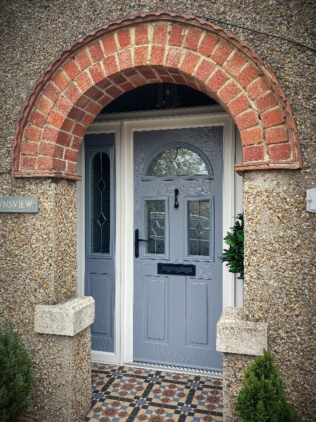 A close-up view of a modern gray composite front door with windows and an arched brick entrance.