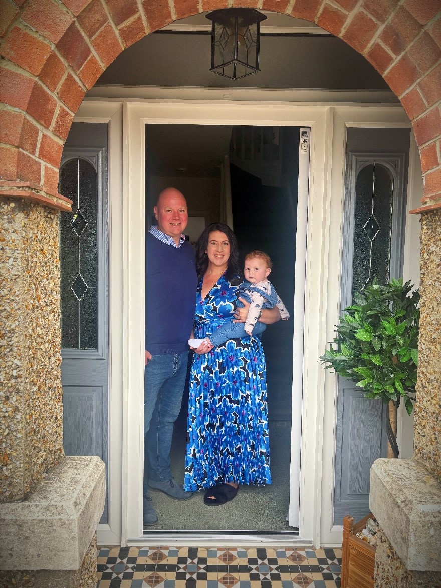 A man, a woman in a blue floral dress holding a baby, standing in a doorway with a tiled floor and an arched brick entrance.
